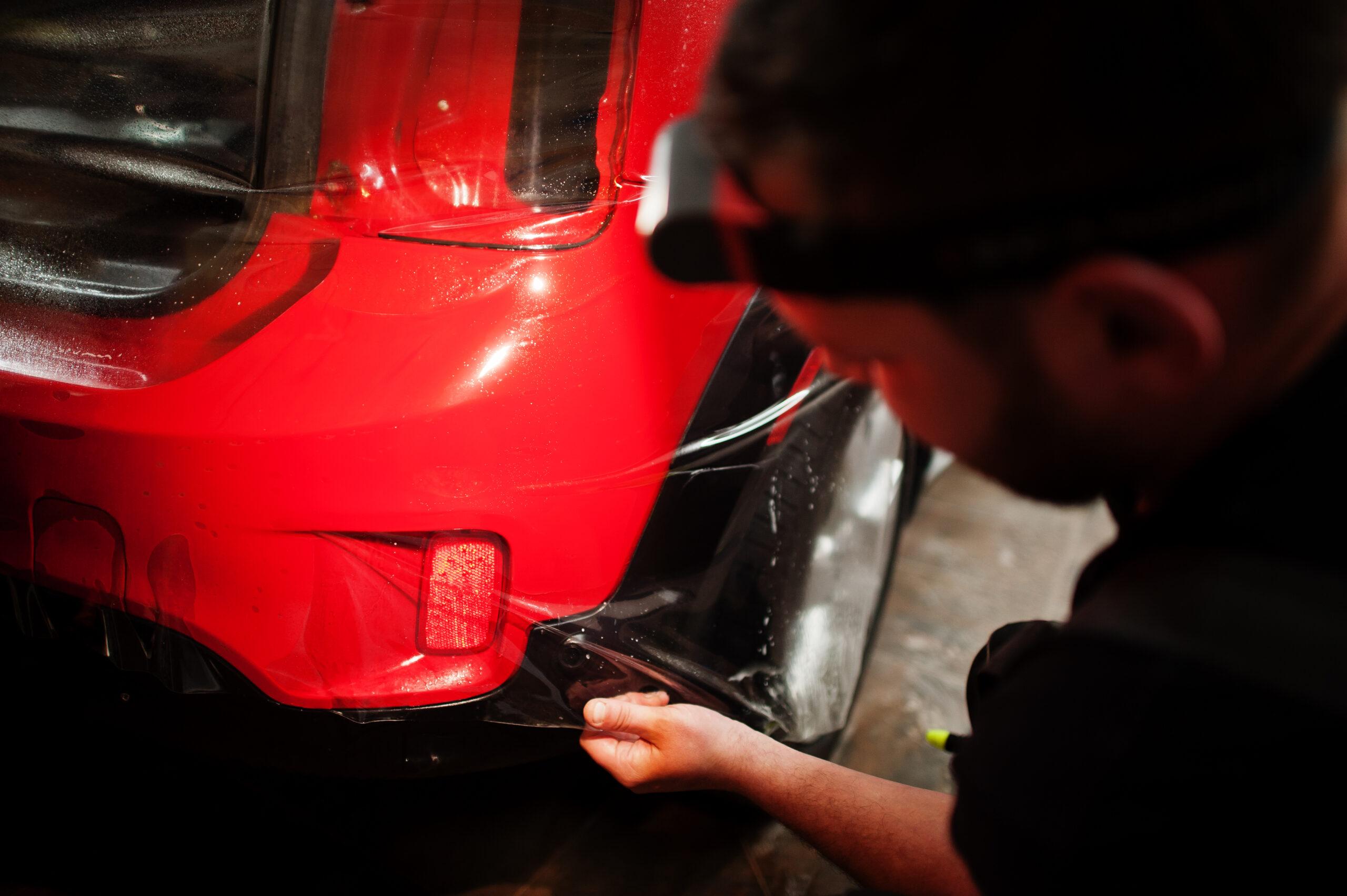 Car service worker put anti gravel film on a red car body at the detailing vehicle workshop. Car protection with special films.